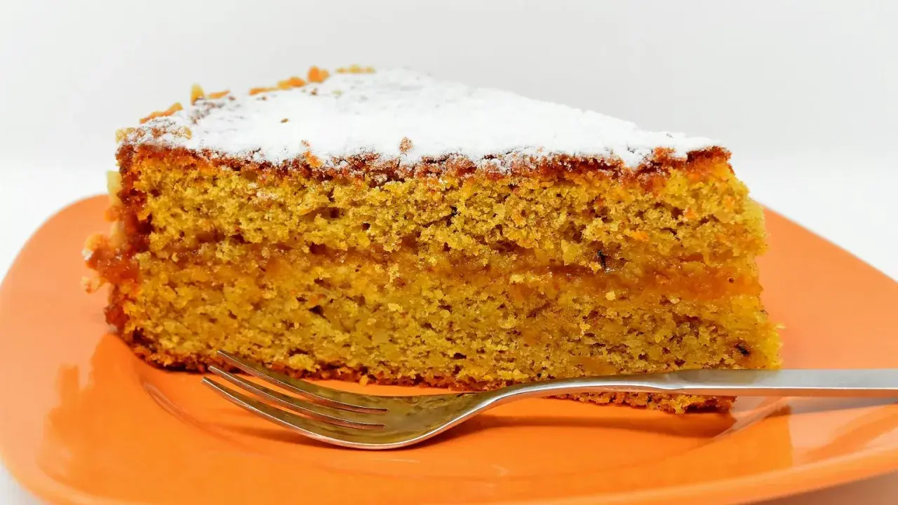 Wilgotne ciasto marchewkowe, posypane cukrem pudrem, idealne jako ciasto dla cukrzyka. Na pomarańczowym talerzu z widelcem.