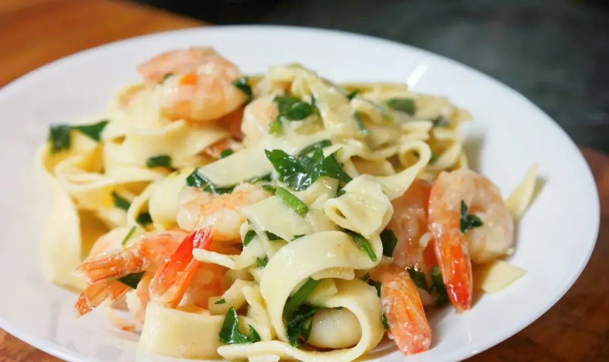 Makaron tagliatelle z krewetkami i zielonymi ziołami w kremowym sosie na białym talerzu.