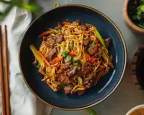 Makaron chow mein z wołowiną i warzywami w misce, obok pałeczek i sosu sojowego.