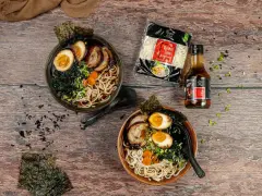 Dwa miski aromatycznego ramen z jajkiem, mięsem i nori. Obok opakowanie makaronu i sosu, idealne składniki, by dowiedzieć się, jak zrobić ramen.