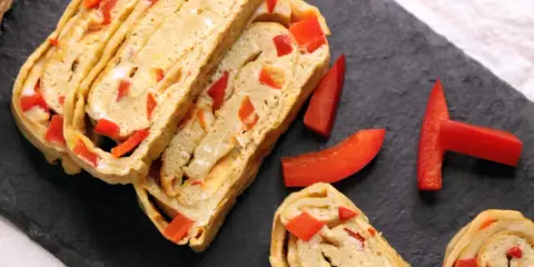Roladki omletu tamago z kawałkami czerwonej papryki, ułożone na czarnej desce.