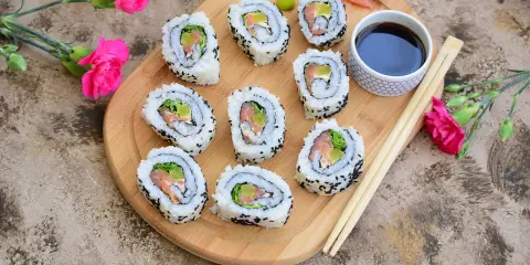Pyszne california sushi z łososiem i awokado, podane na desce z różowymi kwiatami i pałeczkami.