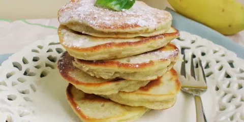 Stos puszystych, trzyskładnikowych placków bananowych, posypanych cukrem pudrem i udekorowanych listkiem, obok dojrzałego banana.