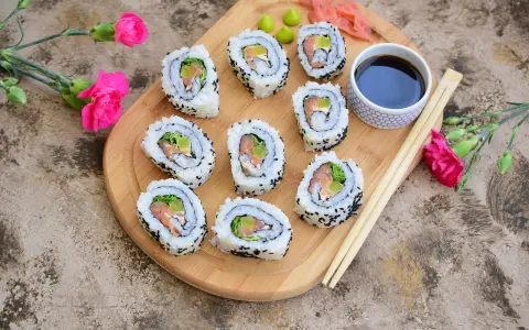 Pyszne california sushi z łososiem i awokado, podane na desce z różowymi kwiatami i pałeczkami.