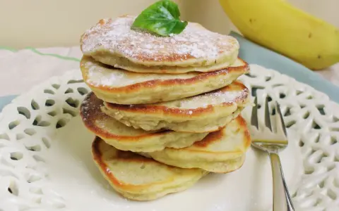 Stos puszystych, trzyskładnikowych placków bananowych, posypanych cukrem pudrem i udekorowanych listkiem, obok dojrzałego banana.