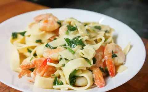 Makaron tagliatelle z krewetkami i zielonymi ziołami w kremowym sosie na białym talerzu.