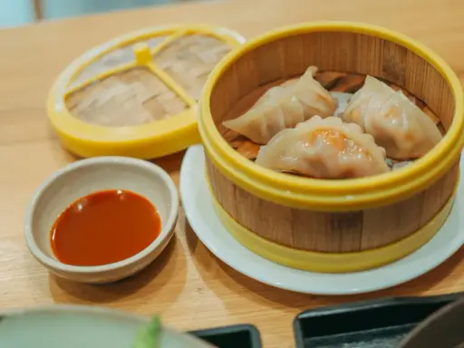 Parujące pierogi gyoza w bambusowym parowniku, obok miseczka z sosem.