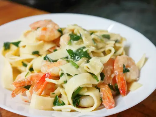 Makaron tagliatelle z krewetkami i zielonymi ziołami w kremowym sosie na białym talerzu.