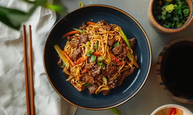Makaron chow mein z wołowiną i warzywami w misce, obok pałeczek i sosu sojowego.
