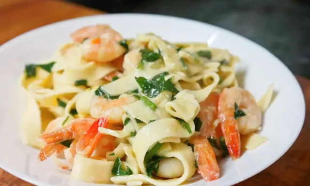 Makaron tagliatelle z krewetkami i zielonymi ziołami w kremowym sosie na białym talerzu.