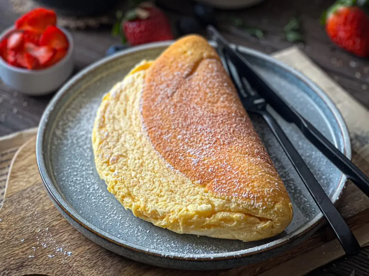 Puszysty omlet jajeczny, lekko przyprószony cukrem pudrem, czeka na talerzu. Obok czarne sztućce i miseczka z truskawkami.
