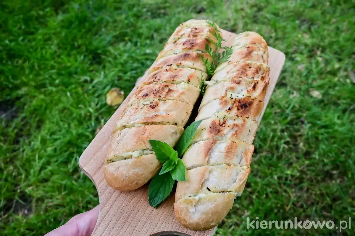 Dwa chrupiące, zapiekane bagietki z ziołami i serem, przypominające pyszne panini, leżą na desce na tle zielonej trawy.