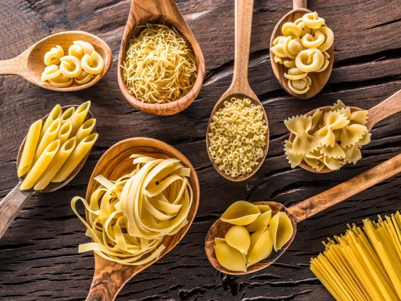 Drewniane łyżki pełne różnorodnego makaronu włoskiego: penne, farfalle, spaghetti, conchiglie i innych.