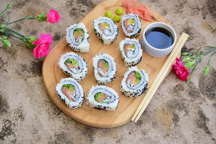 Pyszne california sushi z łososiem i awokado, podane na desce z różowymi kwiatami i pałeczkami.