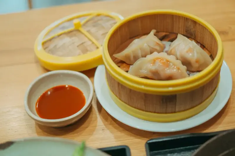 Parujące pierogi gyoza w bambusowym parowniku, obok miseczka z sosem.